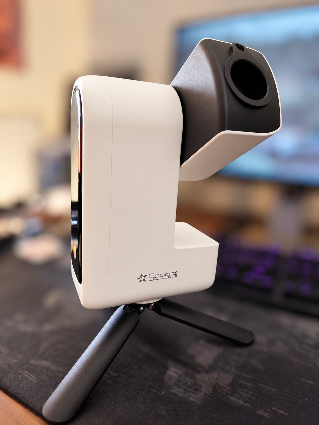 The ZWO Seestar S30 smart telescope on its small tripod: a white body with a dark grey lens housing tilted up, against a softly blurred room.