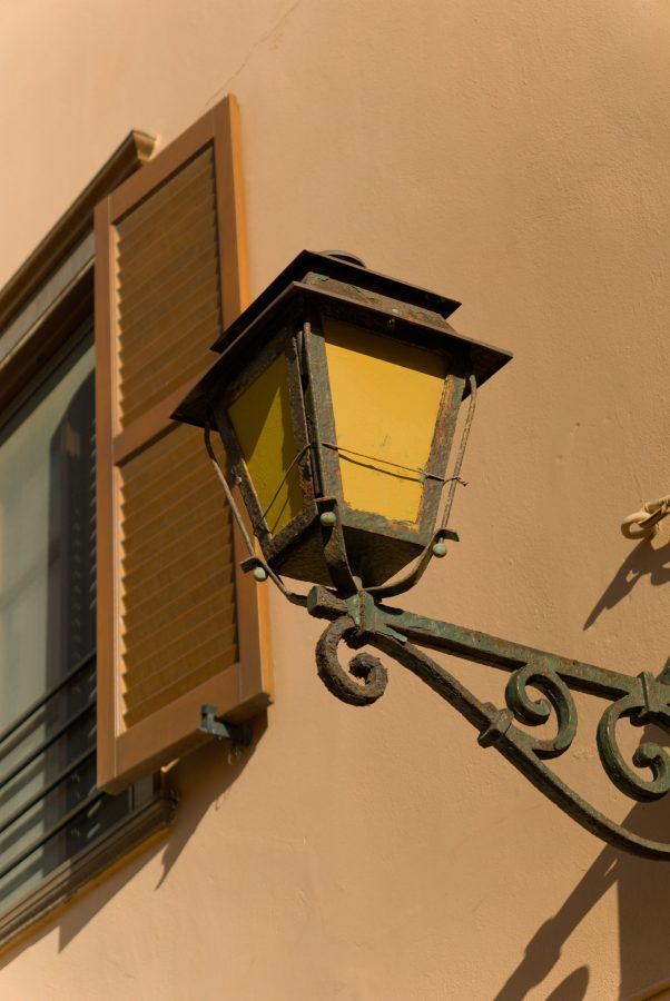 An old wrought-iron street lantern with yellow panes and green, rusting scrollwork on a peach wall, beside a wooden window shutter, in hard afternoon sun.