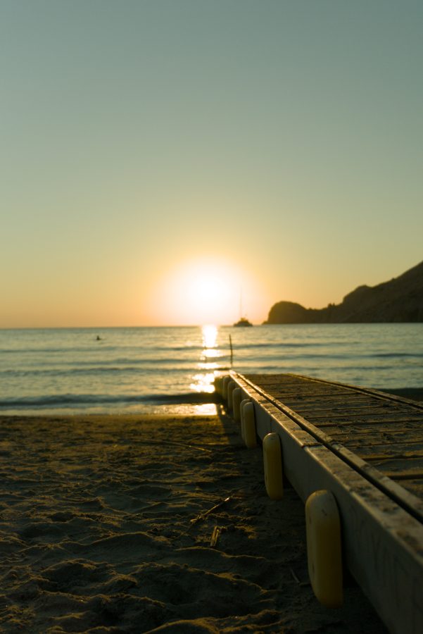 Sunset over a calm sea from beach level: a walkway with yellow bumpers runs into the water, a catamaran sits under the low sun and a headland is silhouetted on the right.