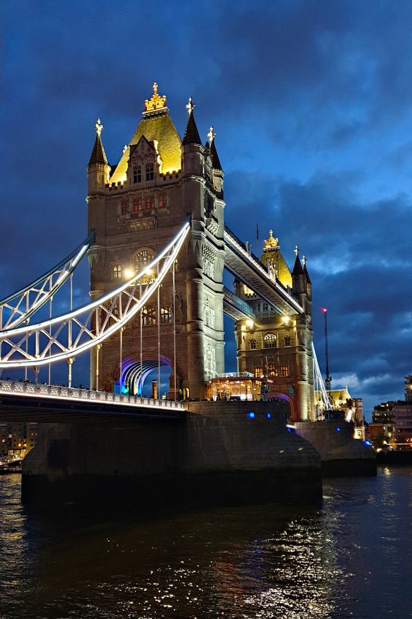 Tower Bridge from the riverside at dusk: its lit Gothic towers with gold pinnacles and pale blue suspension chains under a deep blue, cloudy sky, reflected in the dark Thames.